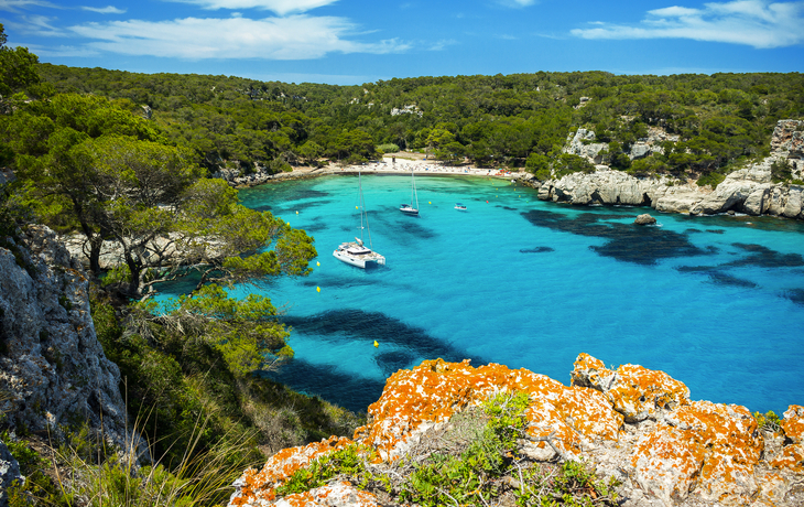 Bucht mit türkisfarbenem Wasser, Segelbooten und bewaldeten Klippen im Hintergrund.