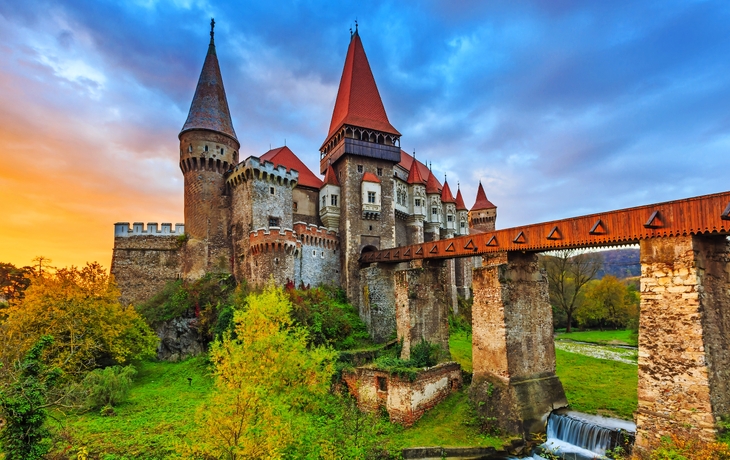Schloss mit roten Dächern bei Sonnenuntergang, umgeben von Bäumen und einem Wasserfall.