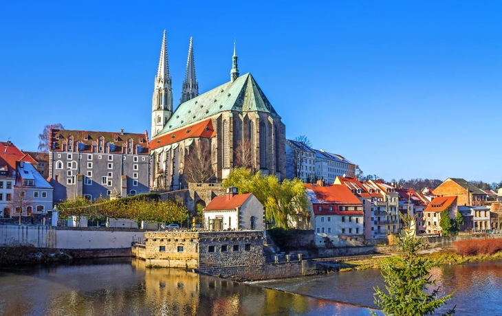 Kirche mit zwei Türmen und roten Dächern am Flussufer unter blauem Himmel.