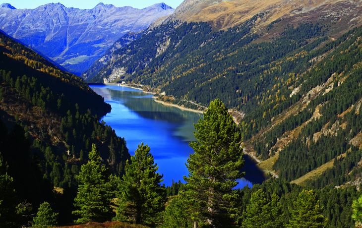 Bergsee umgeben von bewaldeten Bergen unter blauem Himmel.