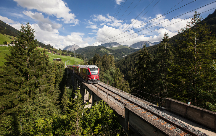 Zug fährt über eine Brücke durch eine bergige Waldlandschaft bei Arosa.