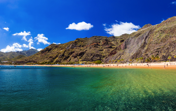 Strand mit türkisfarbenem Wasser und Bergen unter blauem Himmel.