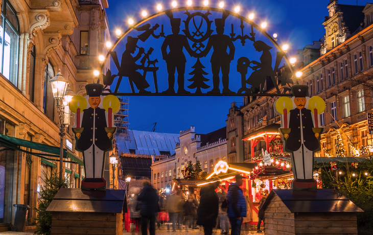 Eingang eines Weihnachtsmarktes mit beleuchtetem Schwibbogen und Menschen.