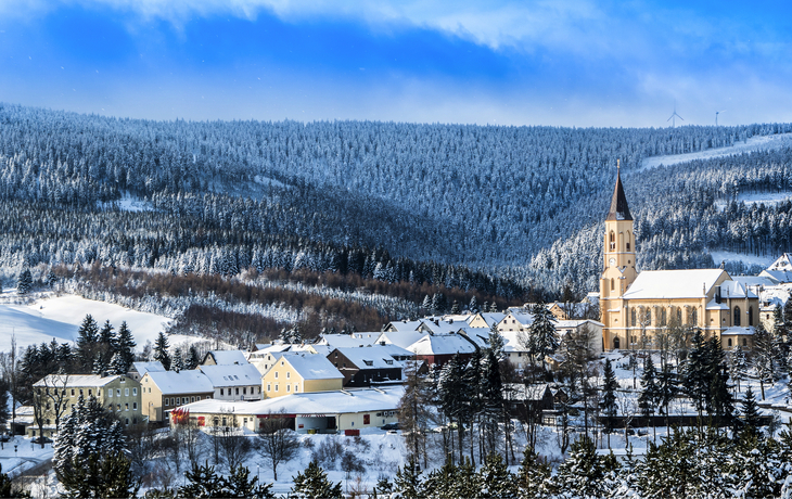 Verschneite Dorfansicht mit Kirche und bewaldeten Hügeln im Hintergrund.