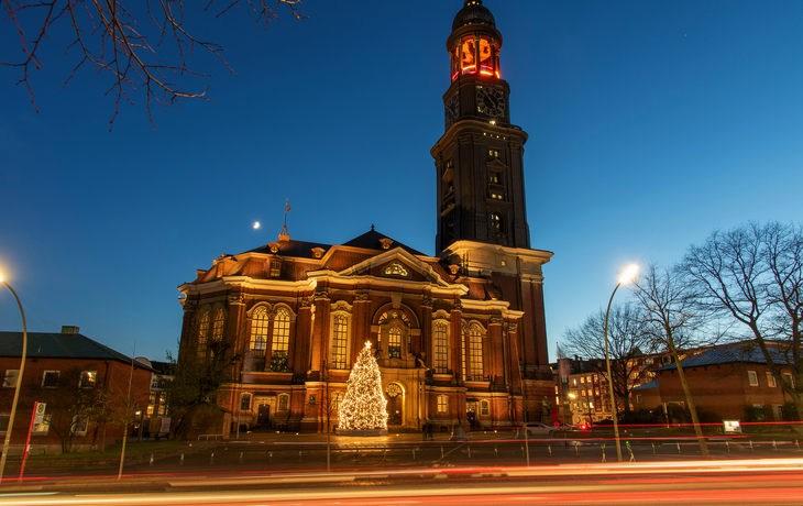 Beleuchtete Kirche bei Dämmerung mit Weihnachtsbaum im Vordergrund.
