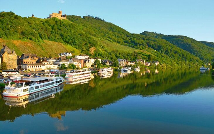 Fluss mit Booten, Häusern und einer Burg auf einem bewaldeten Hügel im Hintergrund.