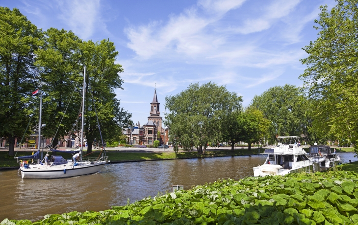 Boote fahren auf einem Fluss an einer Kirche und Bäumen vorbei.