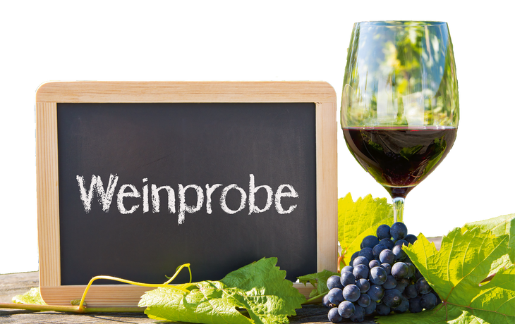 Weinglas und Kreidetafel mit Schriftzug "Weinprobe", umgeben von Weinreben und Trauben.