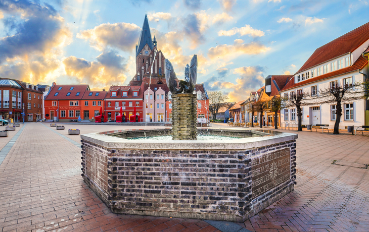 Ein zentraler Platz mit Brunnen und Kirche im Hintergrund bei Sonnenuntergang.