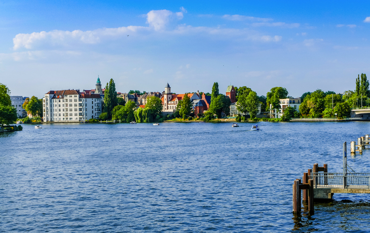 Flusslandschaft mit Uferhäusern und bewölktem Himmel im Hintergrund.