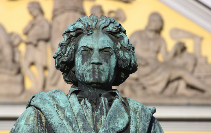 Bronzestatue eines Mannes mit ernstem Gesichtsausdruck vor einem Relief.