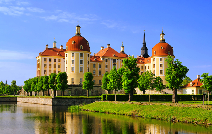 Schloss mit Türmen und rotem Dach am Wasser unter blauem Himmel.