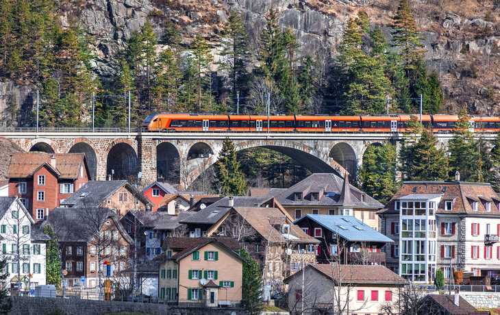 Zug fährt über ein Viadukt in alpiner Landschaft, darunter bunte Häuser.