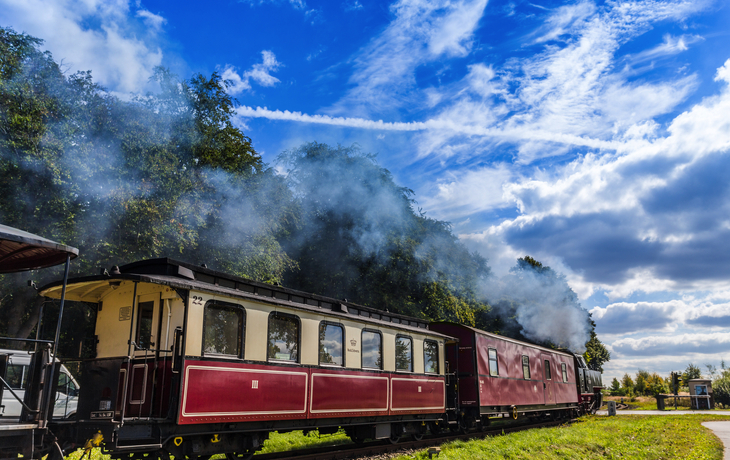 Historische Dampflok mit Dampfwolken vor blauem Himmel und Wolken.
