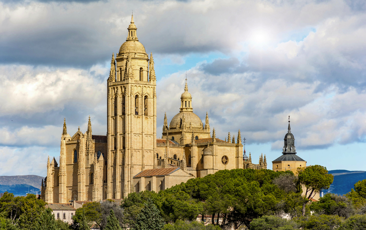Kathedrale von Segovia in Spanien, umgeben von Bäumen, mit bewölktem Himmel im Hintergrund.