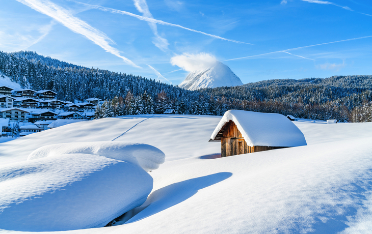 Verschneite Landschaft mit Hütte und Bergen im Hintergrund bei blauem Himmel.