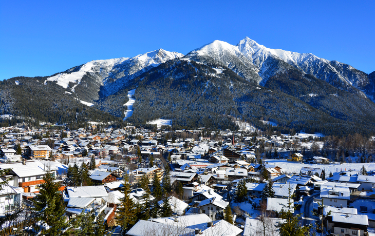 Verschneite Stadt vor schneebedeckten Bergen unter blauem Himmel.