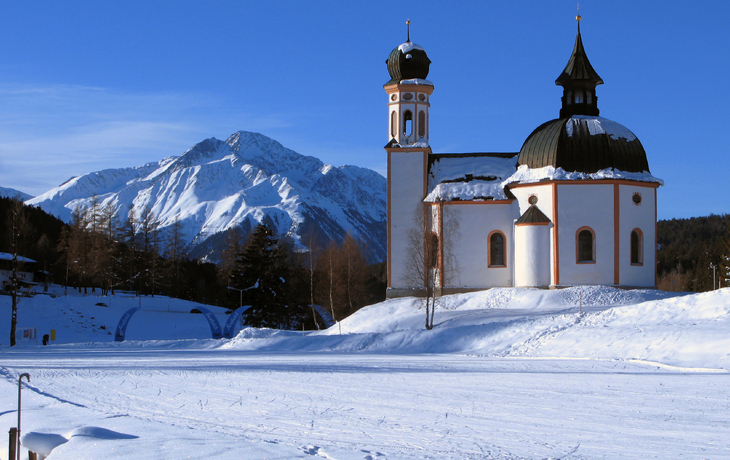 Kirche in verschneiter Alpenlandschaft mit Bergen im Hintergrund.