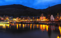 Beleuchtete Stadtansicht am Flussufer bei Nacht mit Hügeln im Hintergrund.